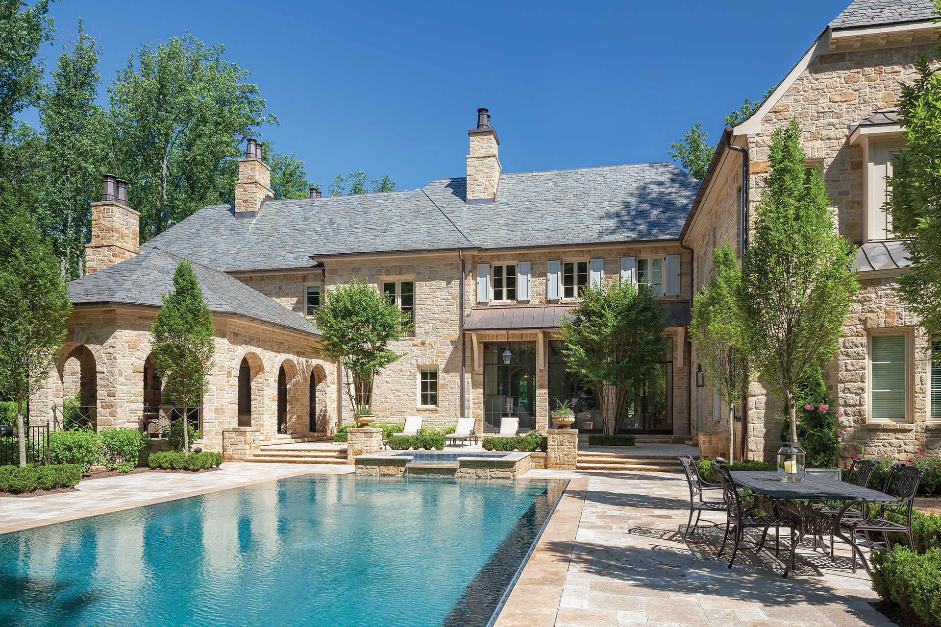 Travertine pool deck is enveloped by boxwood, crape myrtle and European hornbeam trees.